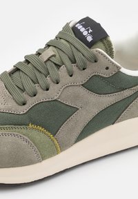 Sneaker verde in una combinazione di suede e tessuto, con un design testurizzato, dettagli di cucitura prominenti e un logo nero sulla linguetta.