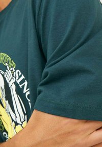 Camiseta de algodón de color verde oscuro con mangas cortas, que presenta un print gráfico de esqueleto en amarillo y blanco en la parte frontal. Textura suave, cuello redondo.