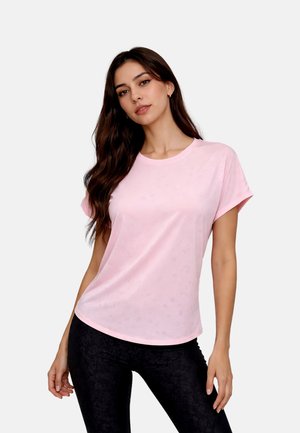 Young woman with long dark hair wearing a light pink short-sleeve t-shirt and black fitted pants, standing against a plain background.