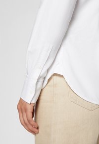 Personne portant une chemise blanche à manches longues rentrée dans un pantalon beige, la main gauche détendue sur le côté.