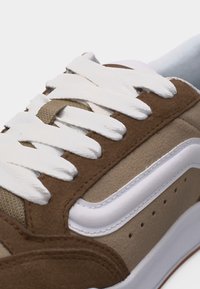 Κοντινό πλάνο καφέ και μπεζ αθλητικού παπουτσιού με λευκά κορδόνια και λευκή ρίγα στο πλάι, που δείχνει λεπτομερή ραφή και υφασμάτινες υφές.