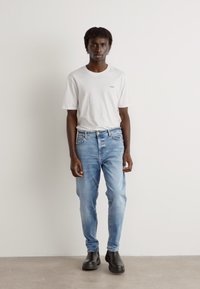Camiseta blanca de cuello redondo, jeans de mezclilla azul claro, botas negras hasta el tobillo. El modelo está de pie contra un fondo blanco liso.