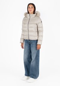 Lichtbeige pufferjack met een bontvoering aan de capuchon, rits aan de voorkant en een gequiltde textuur, gecombineerd met losse blauwe spijkerbroek en witte sneakers.