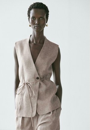 Donna che indossa un gilet beige senza maniche con cintura annodata e pantaloni larghi abbinati, in piedi davanti a uno sfondo chiaro uniforme.