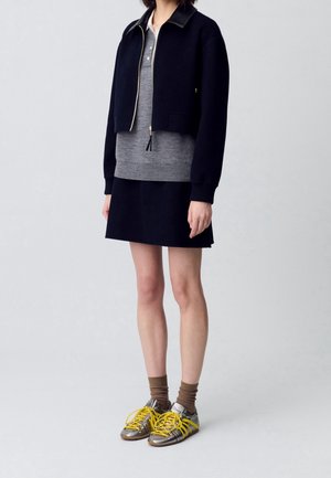 Female model wearing a black cropped zip-up jacket, gray polo shirt, black skirt, brown socks, and metallic silver sneakers with yellow laces.