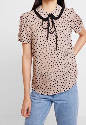 Frau trägt eine beige Kurzarmbluse mit schwarzen Punkten, Rüschenkragen mit schwarzer Schleife und hellblaue Jeans.
