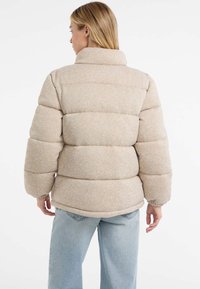 Jachetă puffer bej cu guler înalt, model matlasat și manșete elastice. Fabricată dintr-un material texturat, călduros, cu o croială lejeră.