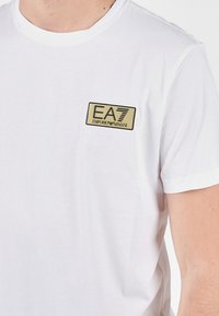 Bílá bavlněná tričko s černým a zlatým logem EA7 Emporio Armani na levé hrudi. Krátké rukávy a kulatý výstřih.