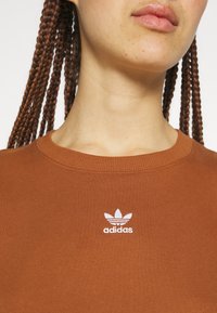 Hnědé bavlněné tričko s žebrovaným límcem a bílým logem Adidas vyšitým na hrudi. Tkanina vypadá hladce a přiléhavě.