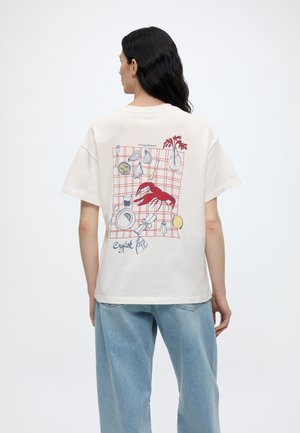 Άτομο που φοράει λευκό μπλουζάκι με κόκκινη απεικόνιση αστακού και θαλασσινών και το κείμενο "Πάρτι Αστακών" στην πλάτη, συνδυασμένο με ανοιχτά μπλε τζιν.