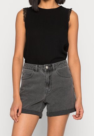 Woman wearing a sleeveless black top with lace trim and high-waisted dark gray rolled-hem denim shorts against a plain background.