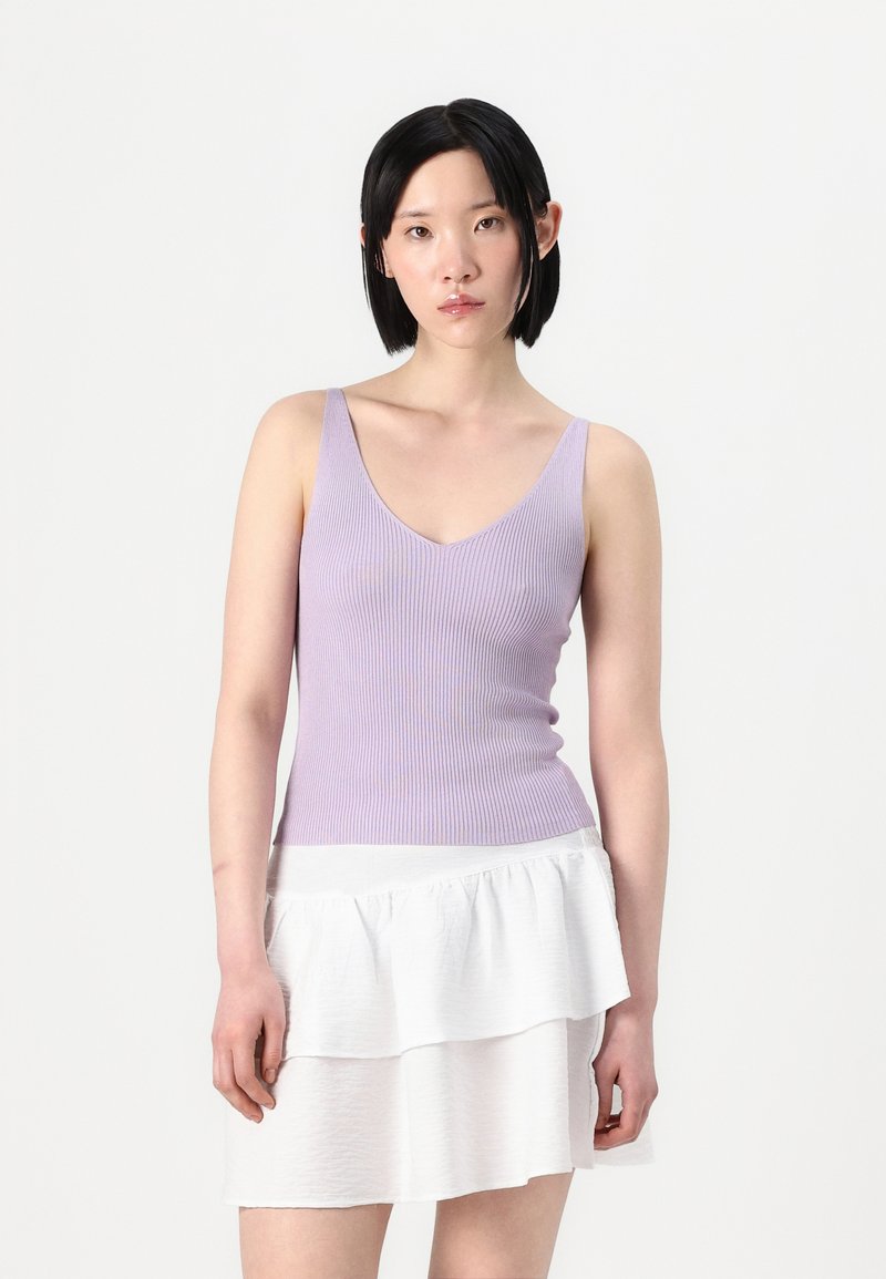 Lys lilla ribbestrikket tanktopp med dyp V-hals, kombinert med en hvit lagdelt mini skjørt med volanger. Glatte teksturer, figursydd design.
