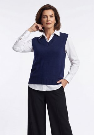 Žena s krátkymi hnedými vlasmi nosí tmavomodrý bezrukávový sveter cez bielu košeľu s golierom a čierne široké nohavice, pózuje s rukou na brade.