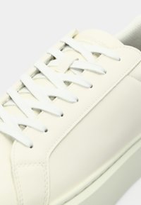 Close-up of a cream-colored sneaker with white laces, smooth leather upper, and a thick rubber sole on a light background.