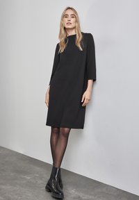 Schwarzes Kleid aus strukturiertem Stoff, runder Halsausschnitt, Dreiviertelärmel und seitliche Schlitze, kombiniert mit Ankle Boots und transparenten Strumpfhosen.
