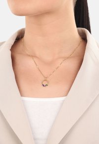 Guldtonad halsband med cirkulär hängsmycke som har lila och gröna detaljer. Bärs över en vit topp, i kombination med en ljusbeige kavaj.