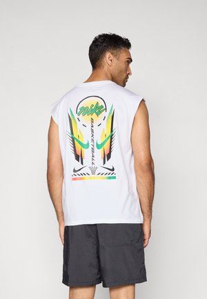 Homme portant un débardeur blanc avec un graphique de basket-ball coloré Nike dans le dos, associé à un short noir, se tenant devant un fond uni.