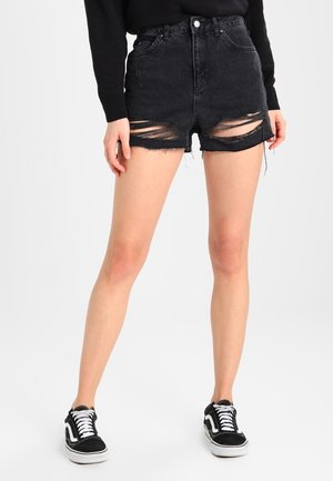 Personne portant un short en jean noir déchiré, un haut noir à manches longues et des baskets noires et blanches, debout sur un fond blanc.