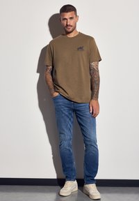 Olivově zelené bavlněné tričko s malým černým logem, spárované s modrými džíny a světle šedými semišovými teniskami, stojící proti jednoduché zdi.