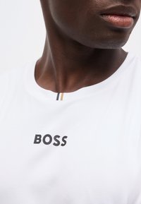 Κοντινό πλάνο ατόμου που φοράει λευκό πουκάμισο με την επιγραφή «BOSS» μαύρα στο στήθος και δύο μικρές κάθετες ρίγες κοντά στη γραμμή του λαιμού.