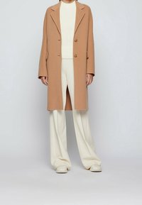 Cappotto lungo marrone chiaro con revers, due bottoni e tasche laterali, indossato sopra un maglione crema e pantaloni a gamba larga crema.