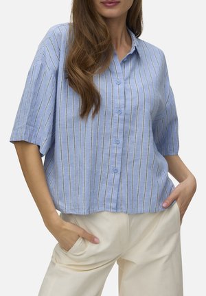 Vrouw die een lichtblauw, kort mouwen overhemd met verticale strepen en crèmekleurige broek draagt met handen in de zakken.