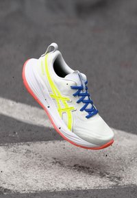 Baskets de course blanches avec des rayures jaunes, lacets bleus et semelle orange, suspendues en l'air au-dessus d'une surface en asphalte.
