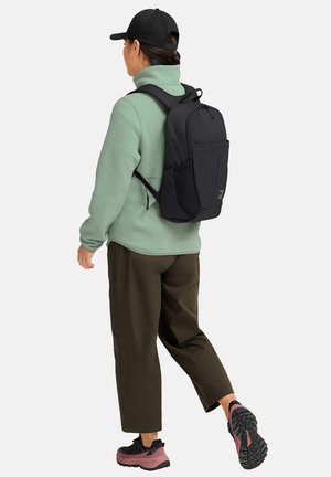 Persona che indossa un cappellino nero, giacca in pile verde, pantaloni scuri e scarpe da trekking rosa e nere, che cammina con uno zaino nero.