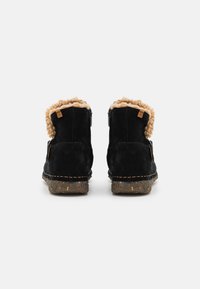 Sorte mockasiner ankelsko med en fluffy tan krage, glidelåslukking, og teksturert brun såle med flerfargede flekker. Synlig sømdetaljering.