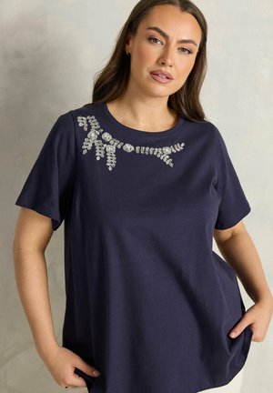 Frau trägt ein marineblaues kurzärmeliges Oberteil mit silbernen Perlenverzierungen in Blumenmuster über der oberen Brust, posiert mit den Händen in den Taschen.