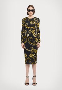 Langermet svart kjole prydet med svingende gule mønstre, med en tettsittende silhuett og kne-lengde kutt. Svart sandaler med stropper fullfører antrekket.