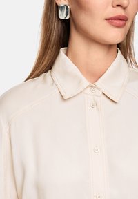 Beige button-up shirt with a classic collar, featuring contrasting stitching, smooth texture, and large rectangular earrings.
