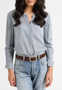 Chemise à manches longues, boutonnée, de couleur bleu clair avec un motif géométrique, associée à un jean bleu délavé et à une ceinture en cuir brun avec une boucle en argent.