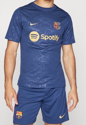 Mężczyzna noszący granatowy sportowy strój FC Barcelona z złotymi logo Nike i Spotify, stojący na jednolitym jasnym tle.