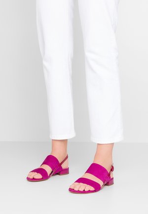 Stopy w jasnoróżowych sandałach na otwartym palcu z dwoma paskami i zapięciem na kostce, w połączeniu z białymi spodniami o długości 3/4, na jednolitym tle.