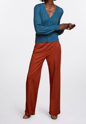 Femme portant un cardigan en maille sarcelle boutonné, un pantalon large couleur rouille avec des boutons sur les côtés, et des chaussures pointues à motif peau de serpent, debout sur fond blanc.