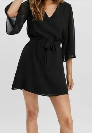 Vrouw draagt een korte zwarte jurk met doorschijnende driekwart mouwen en een ceintuur in de taille, staand met één arm omhoog.