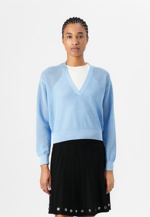 Hellblaues Netzpullover mit V-Ausschnitt und geripptem Saum, kombiniert mit einem schwarzen Faltenrock, der an der Unterkante metallische Ösen aufweist.