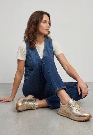 Vrouw met golvend haar die met gekruiste benen op de vloer zit, draagt een wit t-shirt, een blauw denim vest en jeans, en glanzende gouden sneakers.
