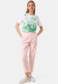 Bluse mit Blumenmuster auf grünem Hintergrund, weiße Ärmel kombiniert mit hellrosa maßgeschneiderten Hosen und weißen Turnschuhen.