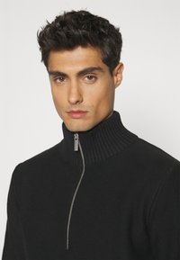Pulover negru tricotat cu guler înalt rânduit și fermoar pe un sfert în față. Material texturat și fermoar argintiu strălucitor adaugă un detaliu elegant.