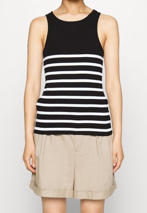 Kobieta ubrana w czarny top bez rękawów z białymi poziomymi paskami i beżowe szorty z podwiniętymi nogawkami, stojąca na jednolitym tle.