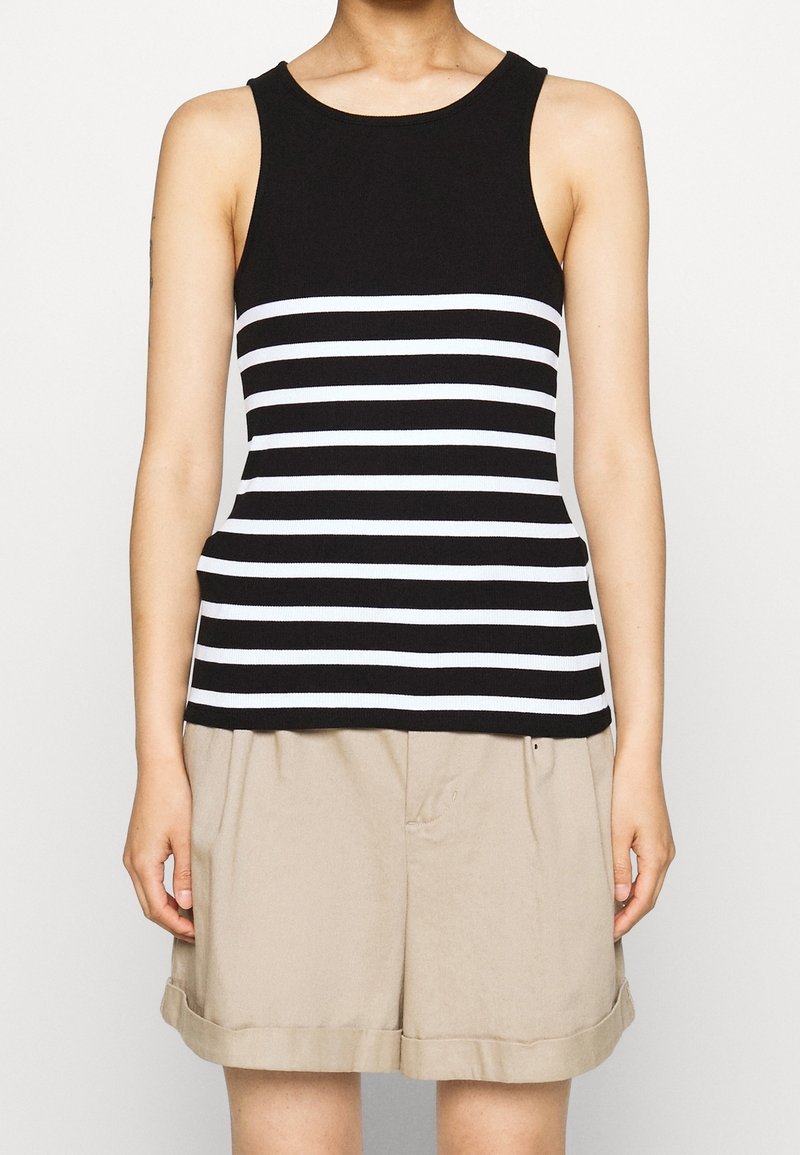 Kobieta ubrana w czarny top bez rękawów z białymi poziomymi paskami i beżowe szorty z podwiniętymi nogawkami, stojąca na jednolitym tle.