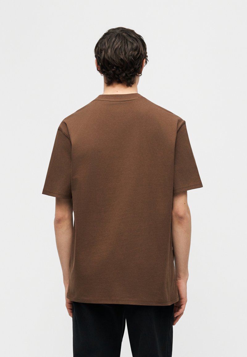 Uomo con capelli scuri che indossa una maglietta marrone semplice a maniche corte, visto di spalle, in piedi contro uno sfondo grigio chiaro.