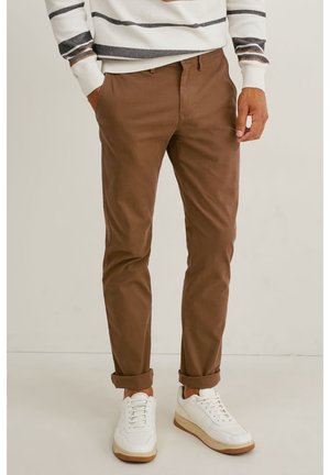 Mężczyzna w brązowych, wąskich spodniach podwiniętych przy kostkach, białych trampkach i białym swetrze z czarno-brązowymi paskami, stojący na jednolitym tle.