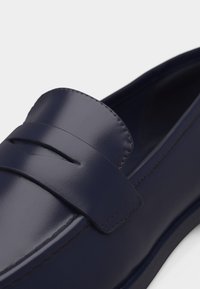 Mocassin en cuir bleu marine avec une finition lisse et polie et un détail de bride sur le dessus, présenté en gros plan.