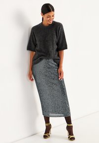 Donkergrijze gebreide trui met korte mouwen, gecombineerd met een maxirok van zilveren pailletten. Model draagt met strappy hoge hakken sandalen.