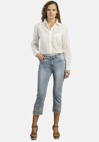 Blusa de algodón blanca bordada con cierre de botones, combinada con pantalones vaqueros cortos de color azul claro con detalles de encaje floral en el dobladillo. Sandalias de plataforma marrones.