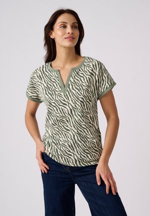 Mujer que lleva una camiseta de manga corta con estampado de cebra y ribete verde, y vaqueros azul oscuro, de pie con una mano en la cadera.