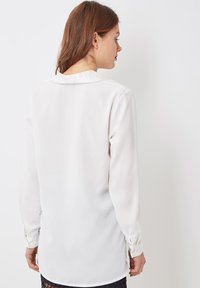 Blusa blanca de manga larga con una textura suave, detalle en el cuello y botones en los puños. La vista trasera muestra un dobladillo ligeramente curvado.